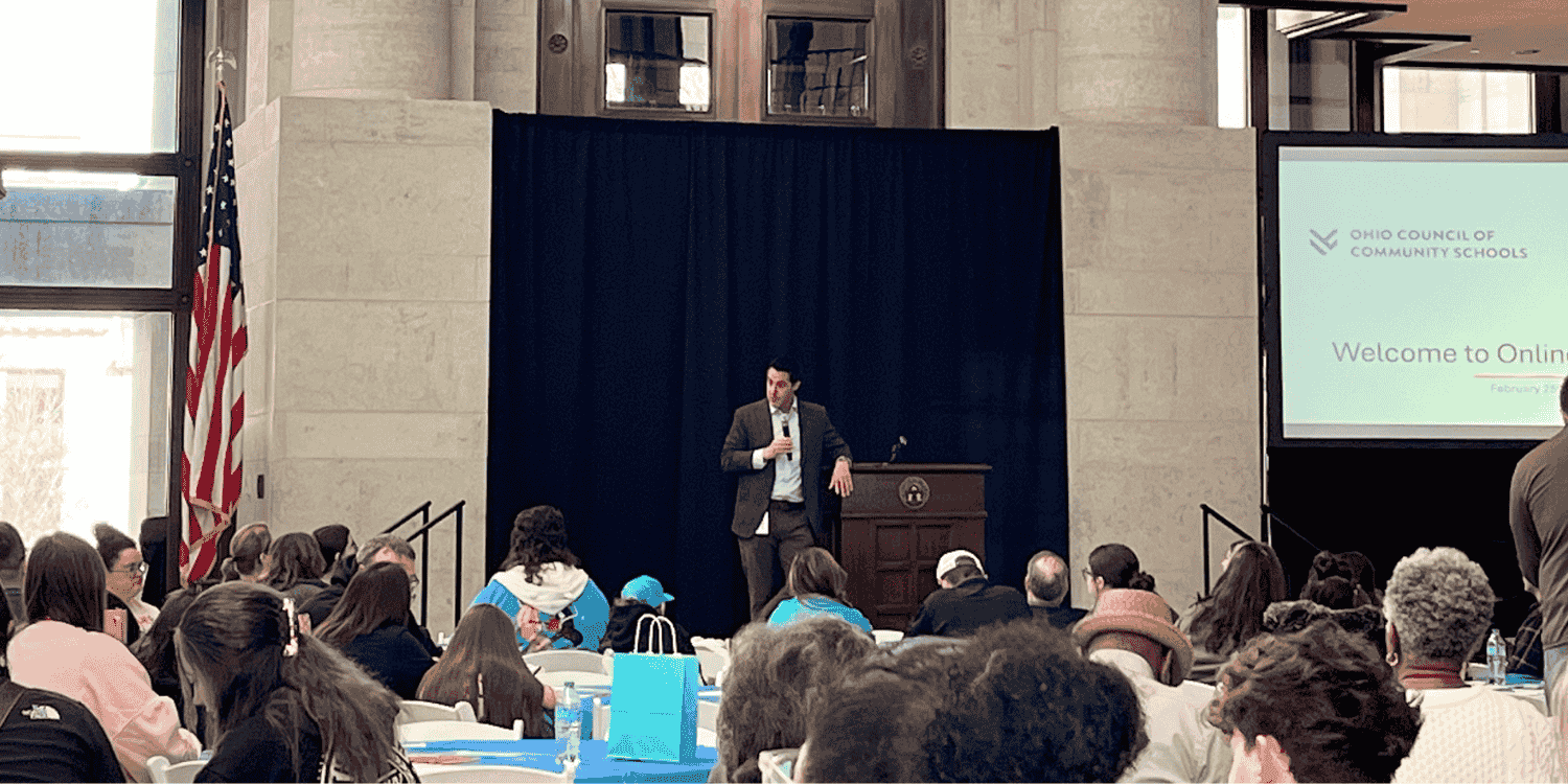 Adult speaker at a podium addresses a seated audience.