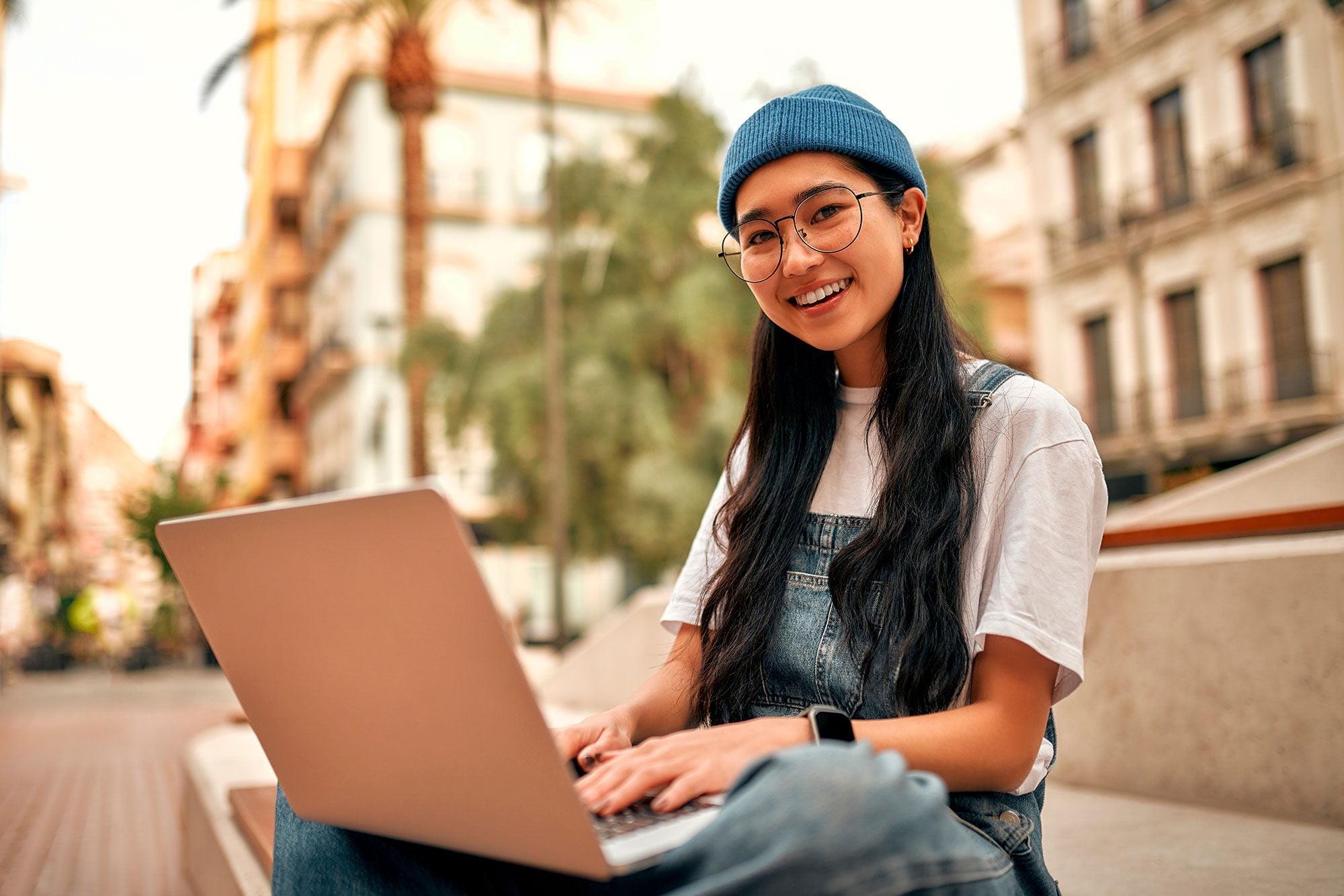 Smiling young adult in beanie and overalls typing on laptop outdoors.